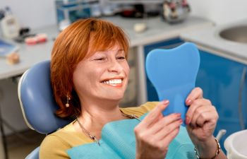 A mature woman after dental restoration treatment sitting in a dental chair and looking at her prefect new smile in a mirror.