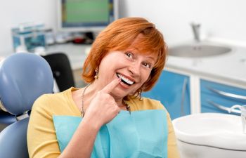 A satisfied mature red-haired woman with a perfect smile sitting in a dental chair and pointing at her tooth.