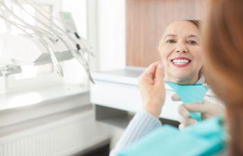 A mature woman after dental implant restoration treatment sitting in a dental chair and looking at her prefect new teeth in a mirror.