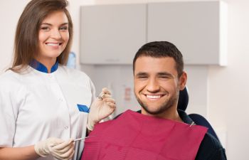 A dentist standing to a happy man with a perfect smile sitting in a dental chair after dental restoration treatment.