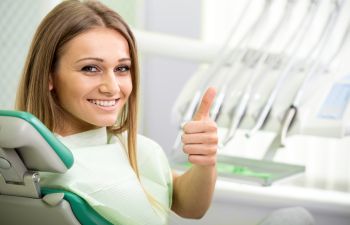 A happy young woman with a perfect smile sitting in a dental chair showing a thumb up.